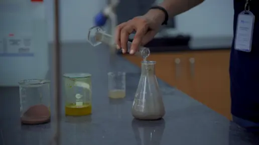 Close-up of a person's hand pouring a liquid into a conical flask on a lab counter