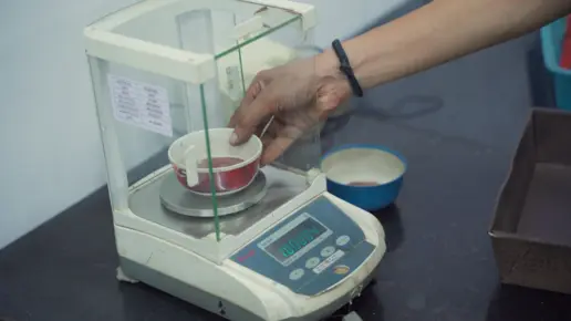 A hand places a cup with red powder on an electronic scale in a lab setting.